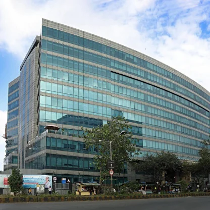 Modern multi story glass office building with a curved facade and reflective blue windows identified as NMIMS University campus, viewed from the street with trees, signage, and a busy roadside in the foreground. The image highlights the contemporary infrastructure and urban location of NMIMS University.